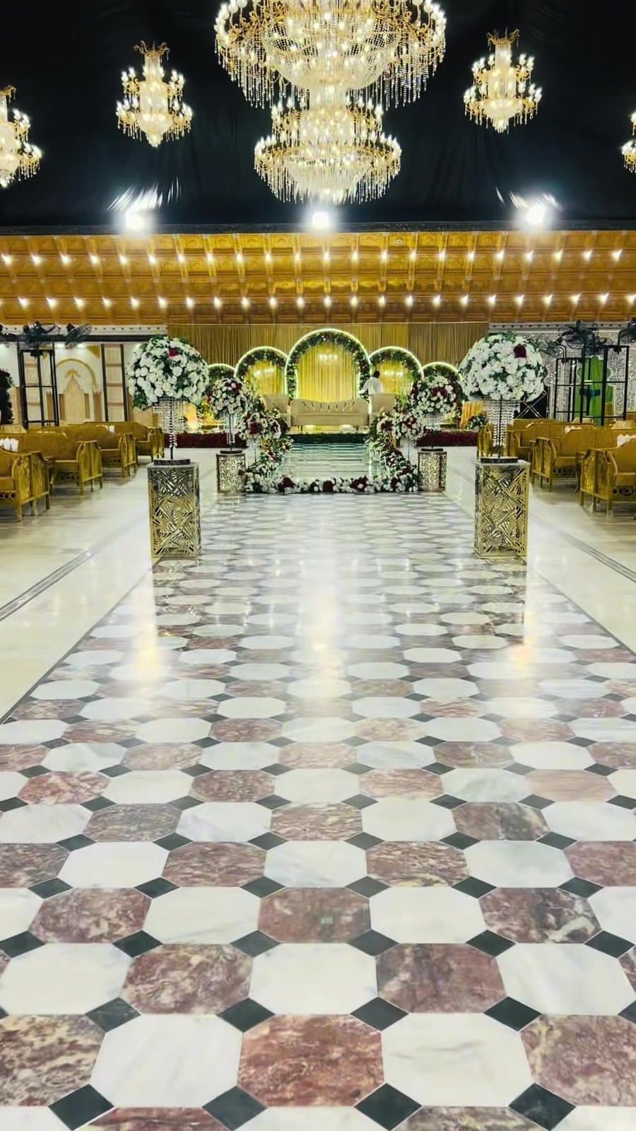 Aisle to the Floral Stage — Crystal chandeliers above a marble aisle leading to an arched floral stage.
